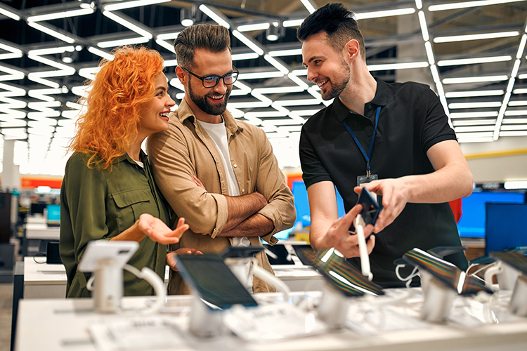 Three people stand at a retail electronics display while a store associate demonstrates a smartphone, with several connected devices arranged on a counter in a modern, well-lit store environment.