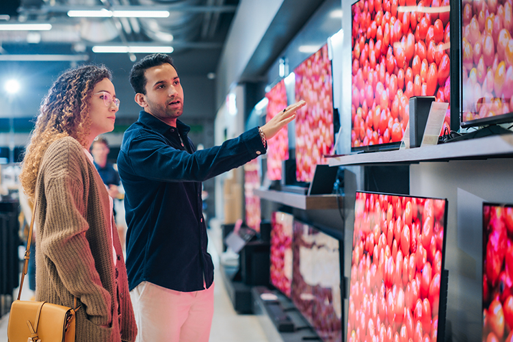 A shopper browses a wall of televisions in an electronics store, with a sales associate pointing toward a display showing vibrant, high-resolution imagery as multiple screens are arranged on sleek shelving in a modern retail environment.