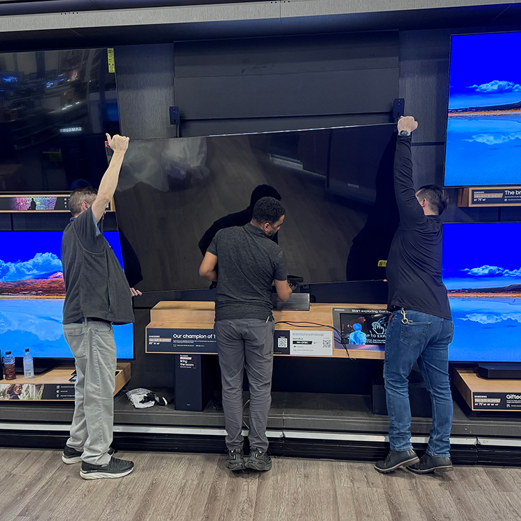 three men work to install a large TV on a display wall at a retail store.